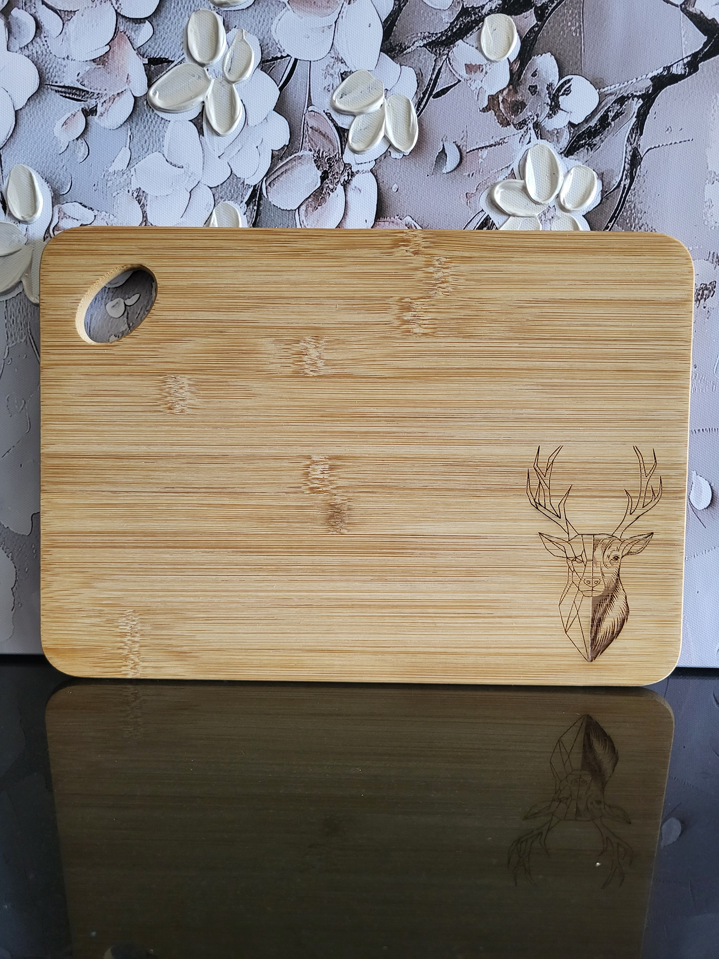 Rechteckiges Schneidebrett aus hellem Bambusholz mit abgerundeten Ecken und seitlicher Grifföffnung. In der rechten unteren Ecke ist eine feine Gravur eines Hirschkopfes mit prächtigem Geweih eingearbeitet, teils im realistischen, teils im geometrischen Stil. Stilvolles und funktionales Küchen- und Servierbrett von ALPCUT – nachhaltig, robust und ideal als Geschenk oder für den täglichen Gebrauch.