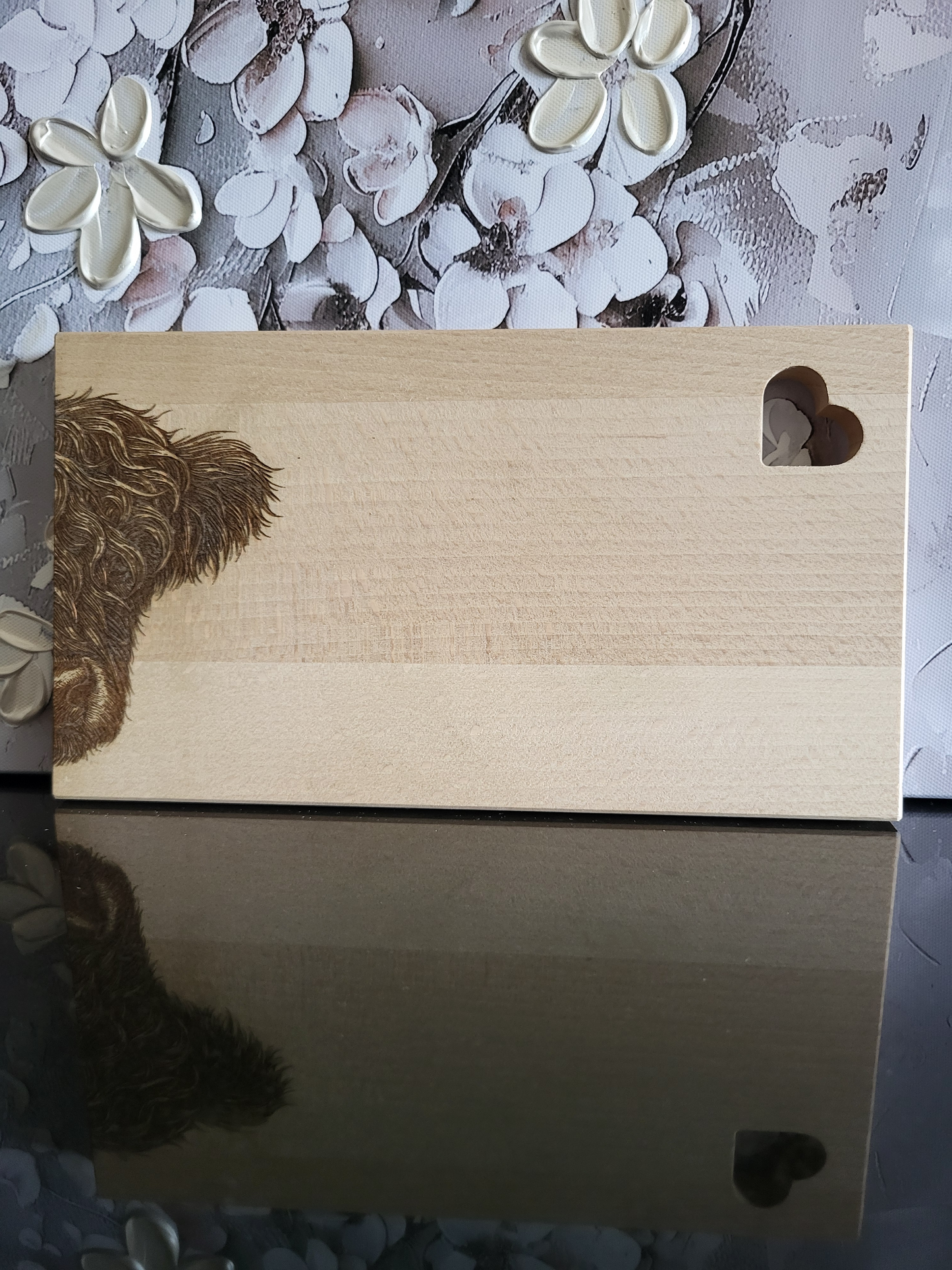 Rechteckiges Schneidebrett aus hellem Buchenholz mit dekorativer Gravur. Auf der linken Seite ist der Kopf eines zotteligen Hochlandrinds detailreich eingraviert, wobei das dichte Fell besonders lebendig wirkt. Rechts oben befindet sich eine ausgestanzte Herzform als praktisches Griffloch, das dem Brett zusätzlich einen charmanten Akzent verleiht. Das massive Holzbrett kombiniert rustikale Natürlichkeit mit liebevollen Details und eignet sich ideal zum Servieren, Schneiden oder als originelles Geschenk.