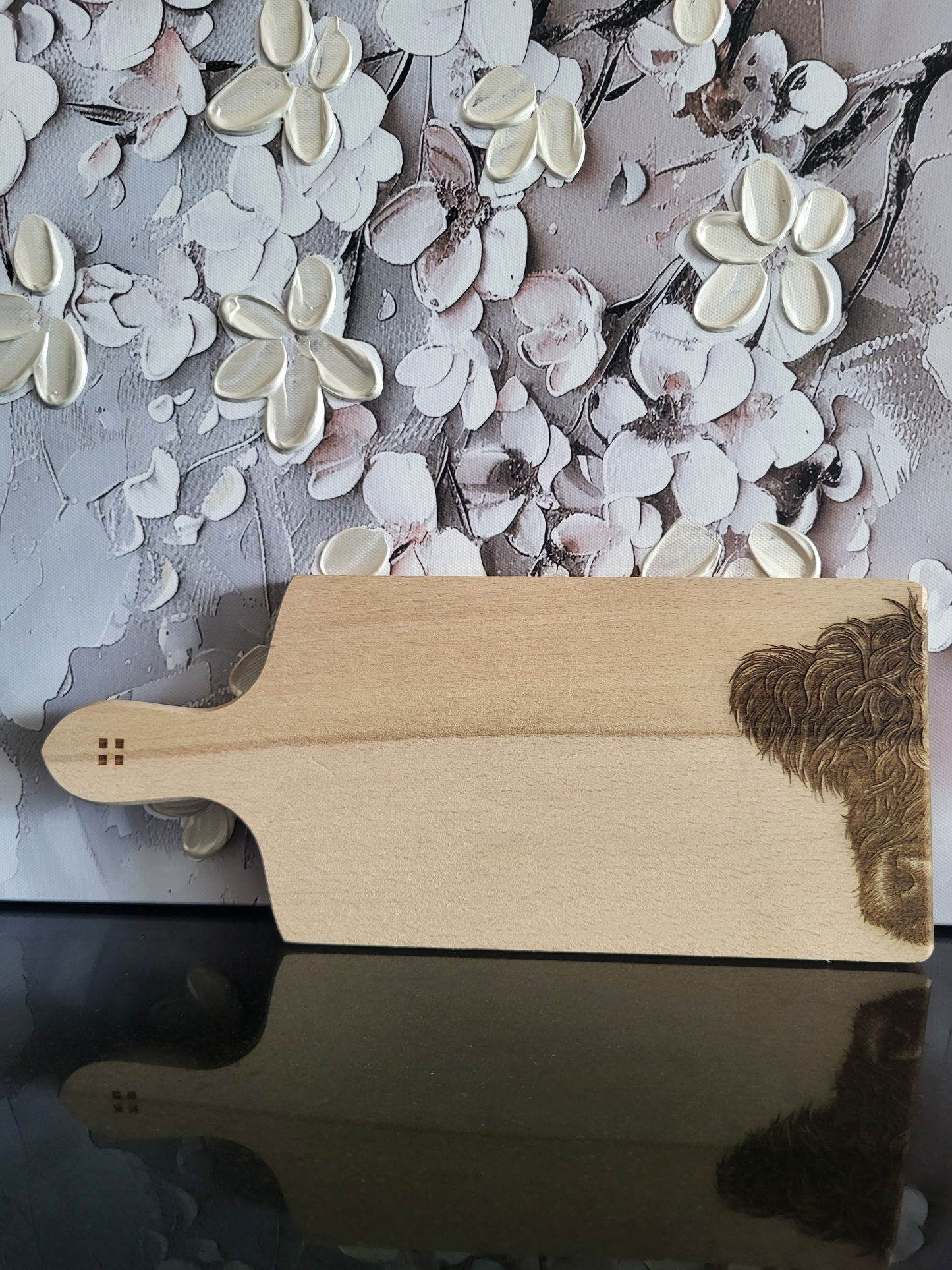 Rechteckiges Schneidebrett aus hellem Buchenholz mit Griff, schlicht und naturbelassen verarbeitet. Auf der rechten unteren Ecke ist detailreich der Kopf eines Hochlandrinds eingraviert, dessen zotteliges Fell besonders plastisch wirkt. Am Griffende befinden sich vier kleine quadratische Einkerbungen als dezentes Design-Element. Das massive Holzbrett vereint Funktionalität mit einem rustikalen, alpinen Charakter und eignet sich ideal zum Schneiden, Servieren oder als Geschenk mit besonderer Gravur.