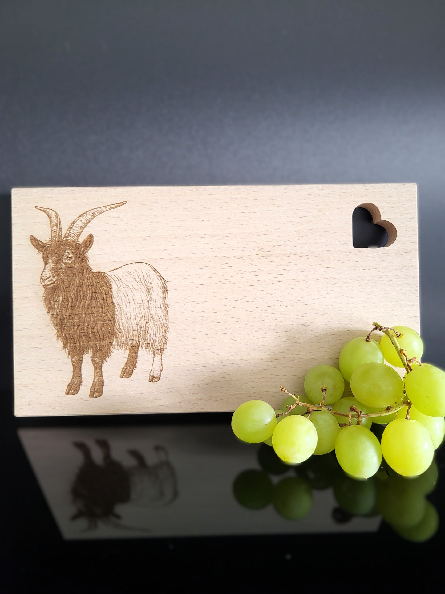 Rechteckiges Schneidebrett aus hellem Buchenholz mit feiner Maserung. Auf der linken Seite ist eine detailreiche Gravur einer Ziege mit langen, geschwungenen Hörnern und zotteligem Fell dargestellt. Auf der rechten oberen Seite befindet sich eine herzförmige Aussparung, die dem Brett einen charmanten Akzent verleiht. Das Brett kombiniert natürliche Schlichtheit mit kunstvoller Gravur – ideal als Küchenaccessoire, Servierbrett oder Geschenk.