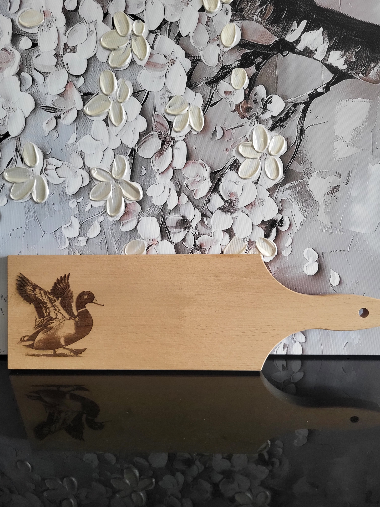 Rechteckiges Schneidebrett aus hellem Buchenholz mit Griff und Loch zum Aufhängen. Auf der linken Seite ist eine detaillierte Gravur einer Ente zu sehen, die mit ausgebreiteten Flügeln im Begriff ist, zu landen oder loszufliegen. Die feine Gravur hebt die Struktur der Federn und den lebendigen Ausdruck hervor. Das Brett kombiniert praktische Funktionalität mit dekorativer Tiergravur und eignet sich sowohl als Küchenutensil als auch als originelles Geschenk.