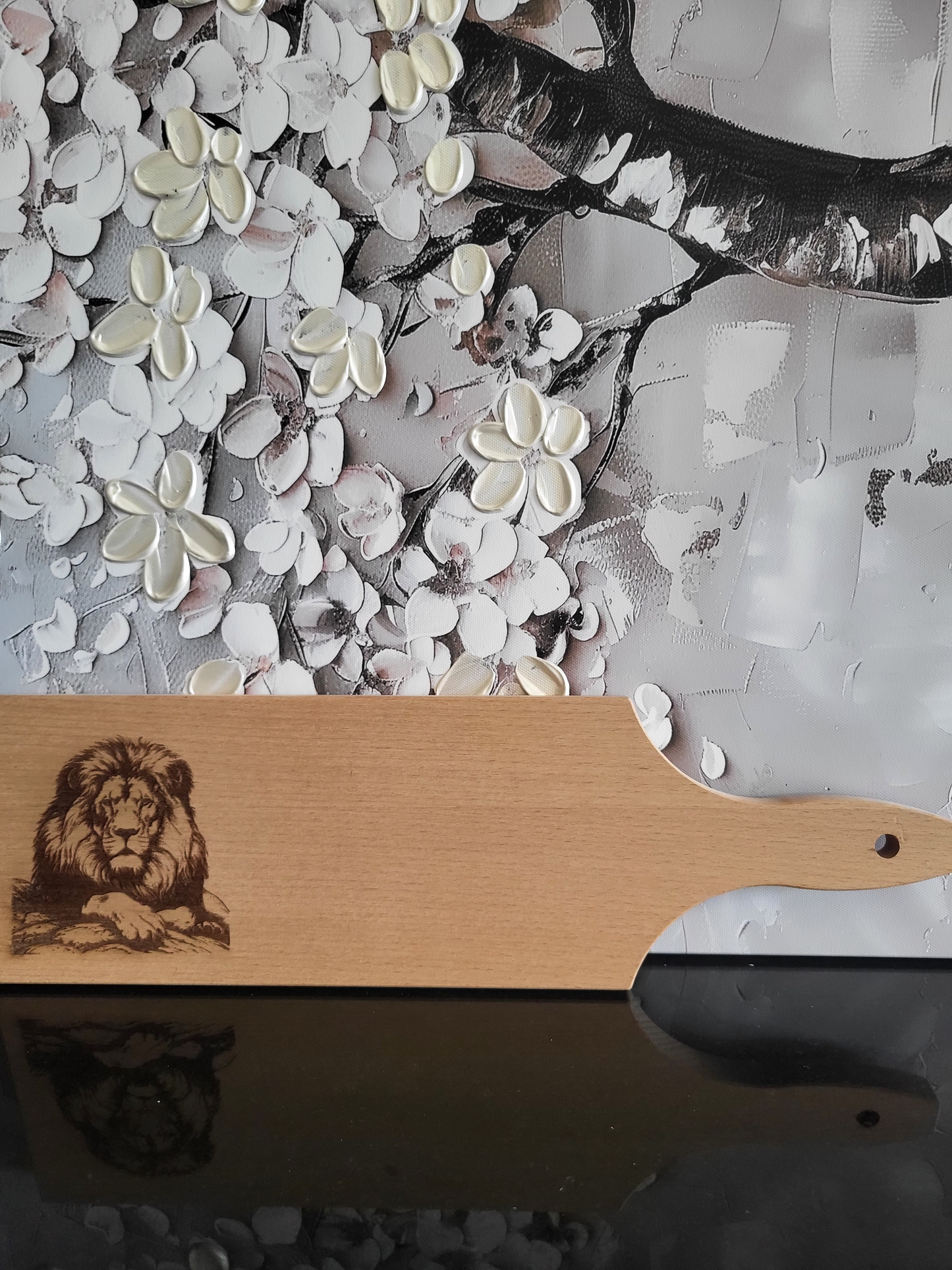 Längliches Schneidebrett aus hellem Buchenholz mit Griff und Loch zum Aufhängen. Auf der linken Seite ist eine detailreiche Gravur eines Löwen zu sehen, der majestätisch auf einem Felsen liegt und nach vorne blickt. Die Gravur zeigt die Mähne, den markanten Gesichtsausdruck und die kraftvolle Haltung des Tieres. Das Holzbrett kombiniert natürliche Schönheit mit kunstvoller Tierdarstellung und eignet sich als dekoratives wie praktisches Küchenaccessoire.