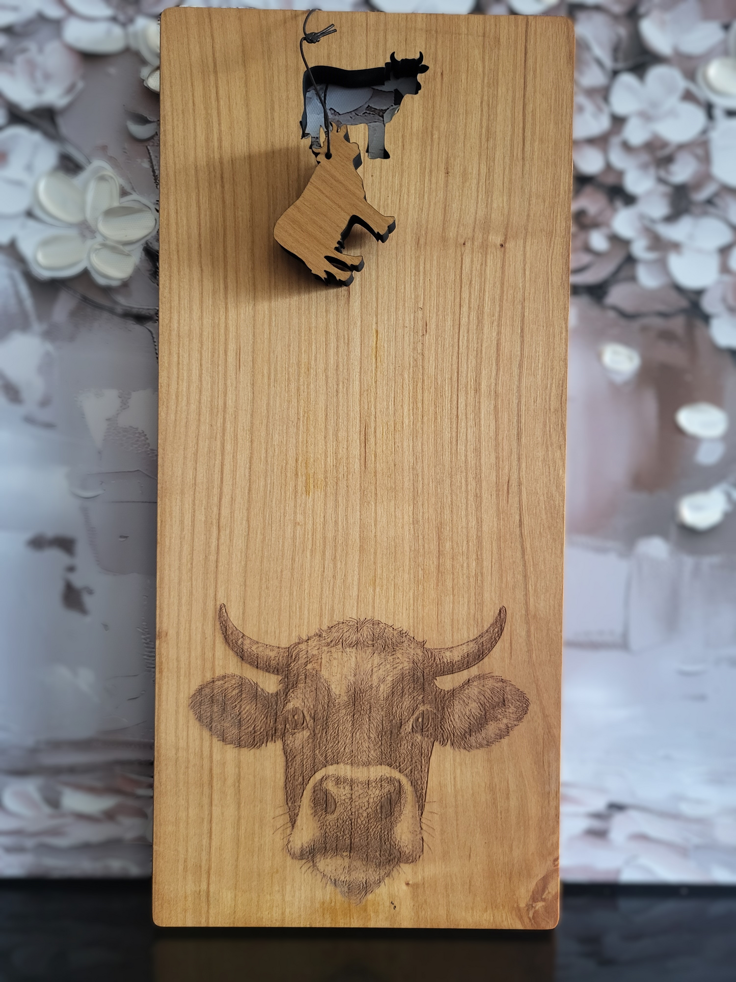 Längliches Schneidebrett aus hellem Holz mit natürlicher Maserung. Am unteren Rand ist detailreich der Kopf einer Kuh mit Hörnern und feiner Fellstruktur eingraviert. Im oberen Bereich befindet sich ein ausgestanztes Kuh-Motiv, aus dem ein kleiner Holzanhänger in Kuhform herabhängt. Das Brett verbindet rustikale Handwerkskunst mit praktischer Funktion und dekorativem Charme.