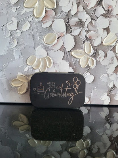 Eine schwarze rechteckige Metalldose, auf deren Deckel eine helle Lasergravur mit dem Schriftzug "Alles Gute zum Geburtstag" zu sehen ist. Daneben sind Symbole wie ein Kuchen mit Kerze und Herzluftballons abgebildet.