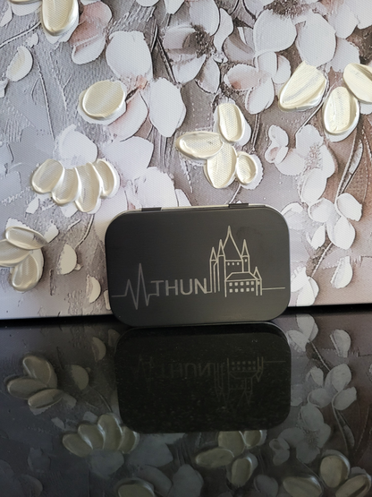 Gravierte Metalldose mit Thun-Motiv. Diese schwarze Box zeigt eine Herzschlag-Grafik mit dem Schriftzug "THUN" und dem berühmten Thuner Schloss. Perfekt als Geschenk oder Souvenir für Reisende, Stadtliebhaber oder Bewohner von Thun. Ideal für die Aufbewahrung von Pillen, Kleingeld oder Schmuck.