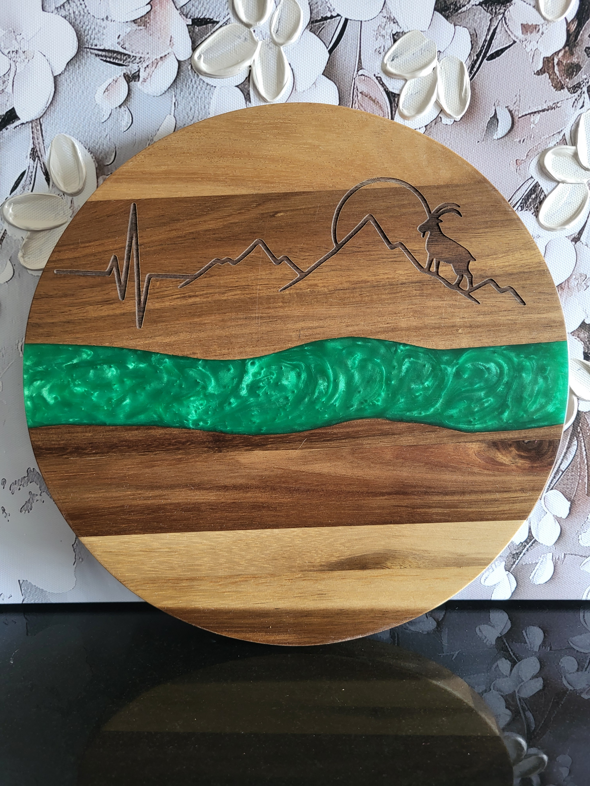 Rundes Schneidebrett aus Akazienholz mit grüner Epoxidharz-Einlage und Gravur eines Steinbocks vor Berglandschaft mit Herzschlaglinie, handgefertigt von ALPCUT – dekoratives Servierbrett und einzigartiges Küchenaccessoire im Alpen-Design, ideal auch als Geschenk.
