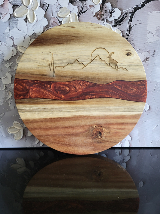 Rundes Schneidebrett aus Akazienholz mit roter Epoxidharz-Einlage und Gravur eines Steinbocks vor Berglandschaft mit Herzschlaglinie, handgefertigt von ALPCUT – edles Servierbrett und dekoratives Küchenaccessoire im Alpenstil, ideal auch als Geschenk für Natur- und Designliebhaber.
