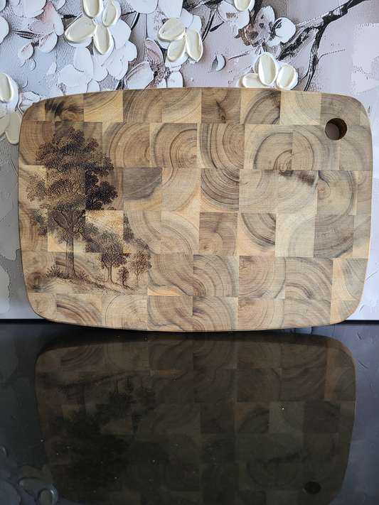 Rechteckiges Schneidebrett aus Akazienholz in Stirnholzoptik mit markanter Blockmusterung. Auf der linken Seite ist eine feine Gravur von Bäumen eingearbeitet, die dem massiven Holzbrett eine natürliche, künstlerische Note verleiht. Hochwertiges, handgefertigtes Küchen- und Servierbrett von ALPCUT – robust, langlebig und ideal zum Schneiden, Servieren oder stilvollen Präsentieren.