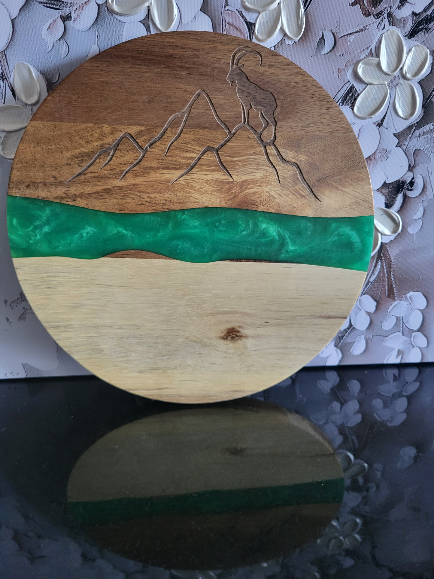 Rundes Brett aus Akazienholz mit Epoxidharz-Band. Die Oberseite hat eine Lasergravur, die eine Berglandschaft mit einem Steinbock darstellt. Ein leuchtend grünes Kunstharz-Band trennt die hellere Unterseite von der dunkleren Oberseite. Ideal als dekoratives Servierbrett für Käse, Wurstwaren oder als Blickfang für Liebhaber von alpinen Motiven.