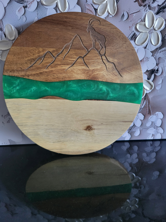 Rundes Brett aus Akazienholz mit Epoxidharz-Band. Die Oberseite hat eine Lasergravur, die eine Berglandschaft mit einem Steinbock darstellt. Ein leuchtend grünes Kunstharz-Band trennt die hellere Unterseite von der dunkleren Oberseite. Ideal als dekoratives Servierbrett für Käse, Wurstwaren oder als Blickfang für Liebhaber von alpinen Motiven.