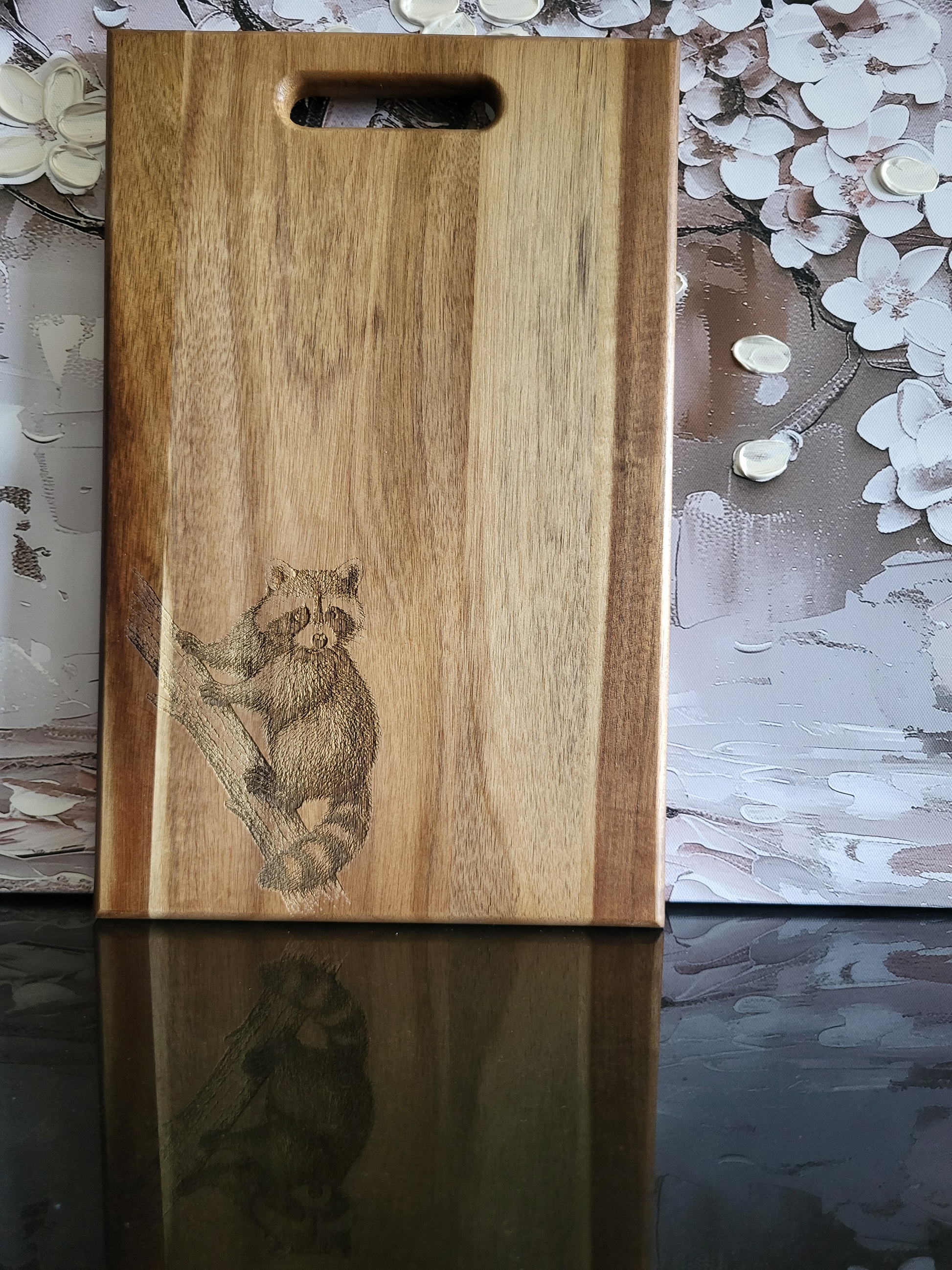 Ein hochformatiges, rechteckiges Schneidebrett oder Holzbrett aus Akazienholz, das oben einen Griff hat. Auf der linken Seite des Brettes ist ein Waschbär abgebildet, der einen Ast hinaufklettert. Das Holz hat eine markante Maserung.