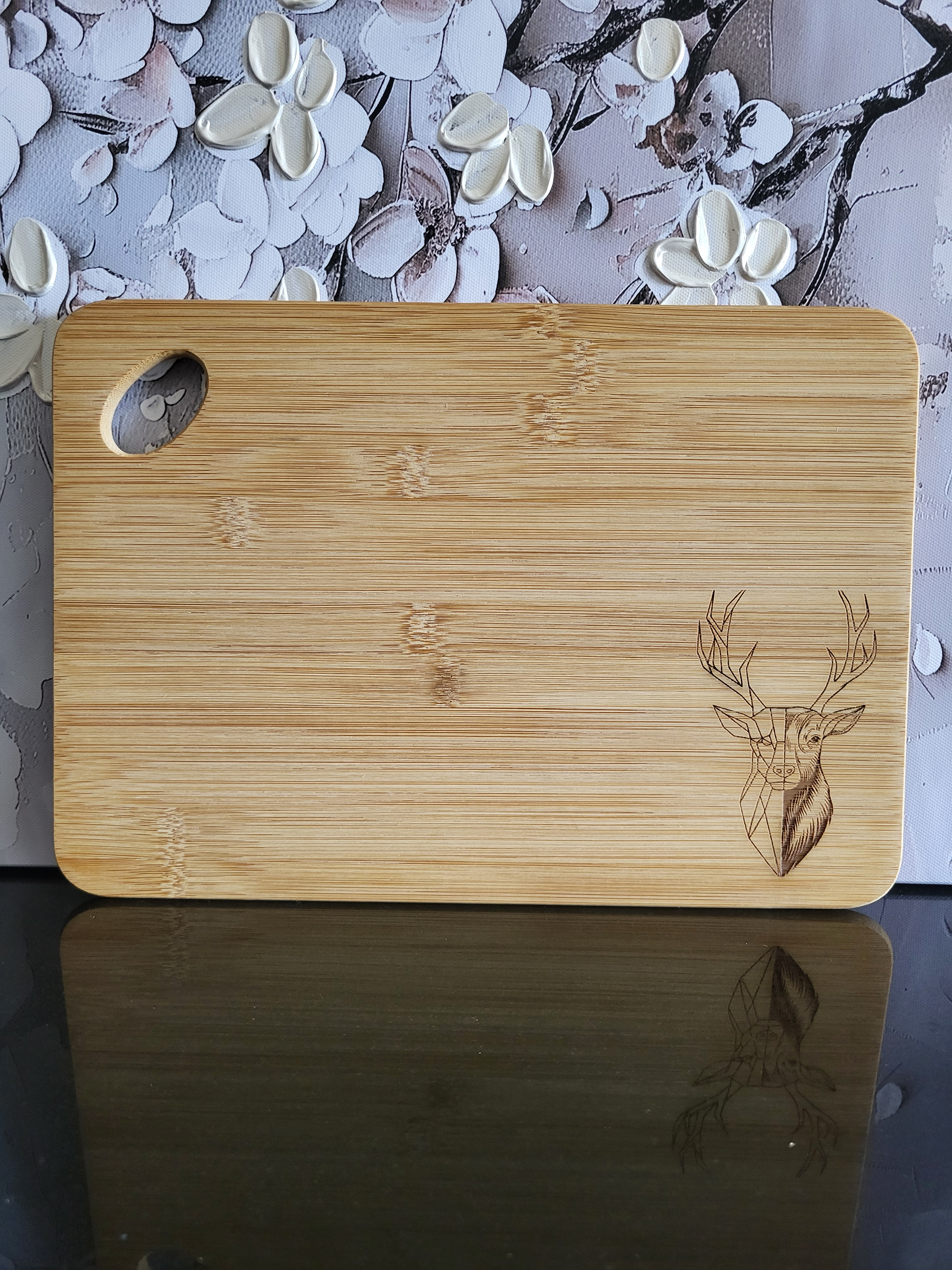 Rechteckiges Schneidebrett aus hellem Bambusholz mit abgerundeten Ecken und seitlicher Grifföffnung. In der rechten unteren Ecke ist eine feine Gravur eines Hirschkopfes mit prächtigem Geweih eingearbeitet, teils im realistischen, teils im geometrischen Stil. Stilvolles und funktionales Küchen- und Servierbrett von ALPCUT – nachhaltig, robust und ideal als Geschenk oder für den täglichen Gebrauch.