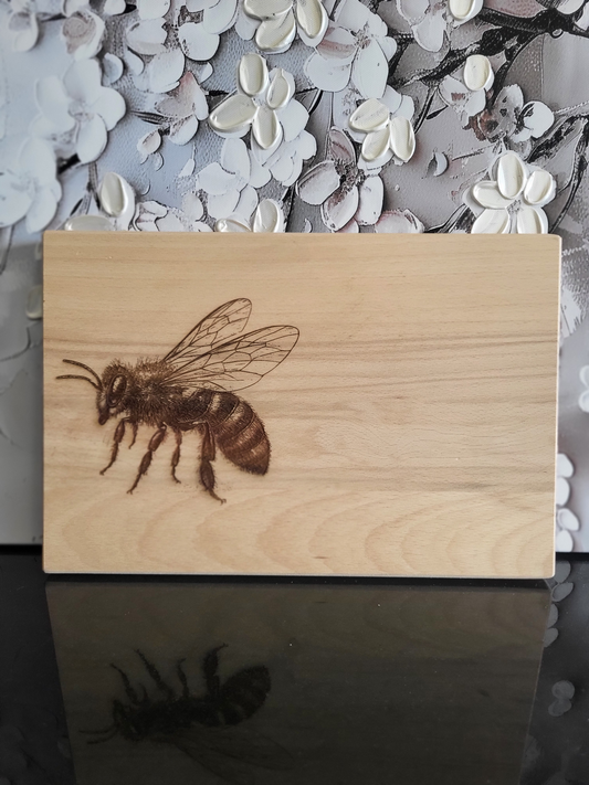 Rechteckiges Schneidebrett aus hellem Holz mit naturbelassener Maserung. Auf der linken Seite ist eine detaillierte Gravur einer Biene zu sehen – fein ausgearbeitet mit erkennbaren Flügelstrukturen, Körpersegmenten und Beinen. Das schlichte Design hebt die Gravur hervor und macht das Brett zu einem stilvollen Küchenaccessoire. Ideal als Schneide- oder Servierbrett nutzbar, verbindet es Funktionalität mit handwerklicher Kunst und Naturverbundenheit.