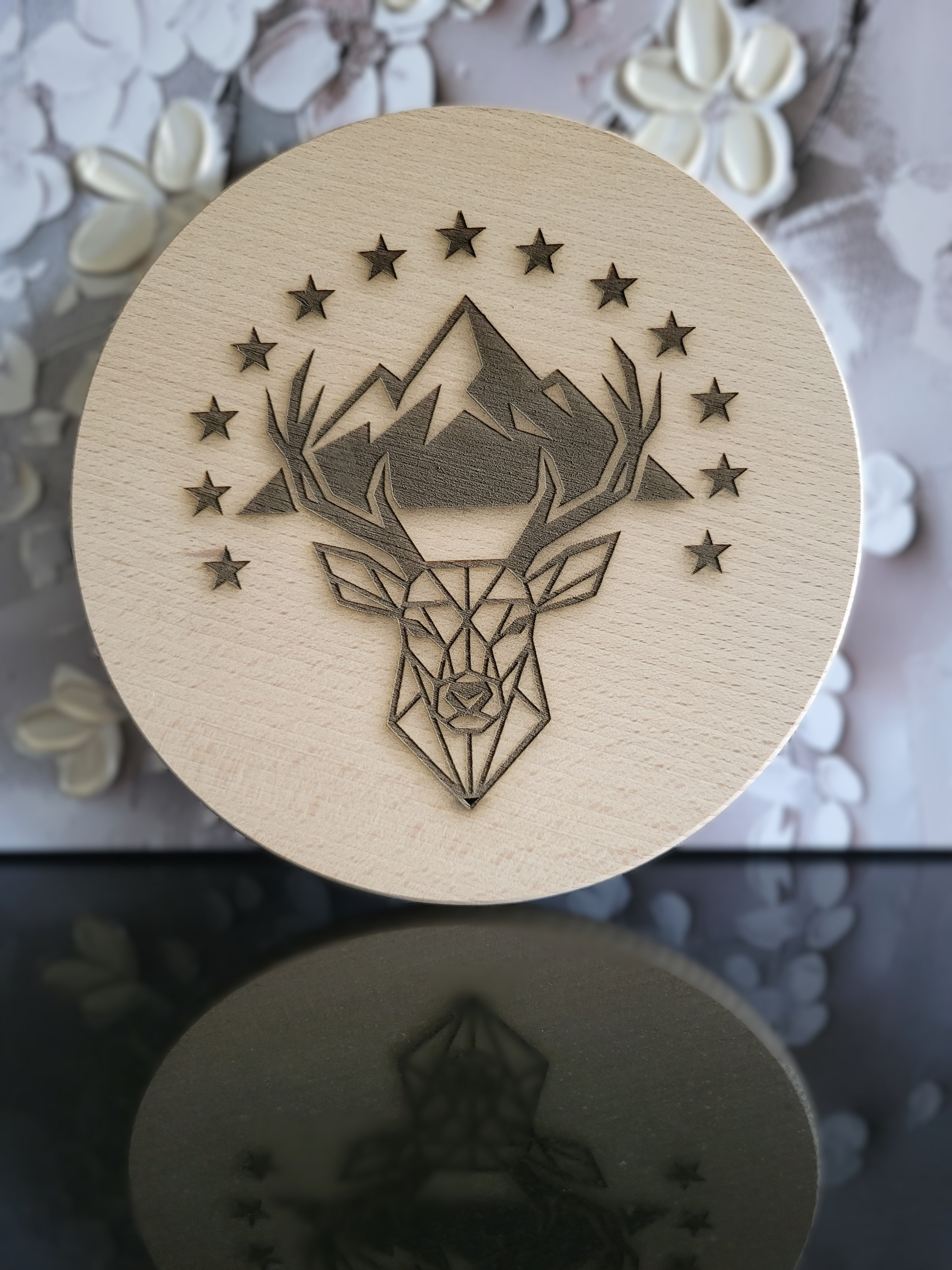Runder Untersetzer aus hellem Buchenholz mit detailreicher Gravur. In der Mitte ist ein geometrisch stilisierter Hirschkopf mit ausladendem Geweih dargestellt. Über dem Hirsch erheben sich Berggipfel, eingerahmt von einem Kreis aus zwölf Sternen. Das naturverbundene Design verbindet alpine Landschaft mit der Symbolkraft des Hirsches und macht den Untersetzer zu einem dekorativen wie praktischen Accessoire – ideal für Tischkultur, Alpenfans und als Geschenk.