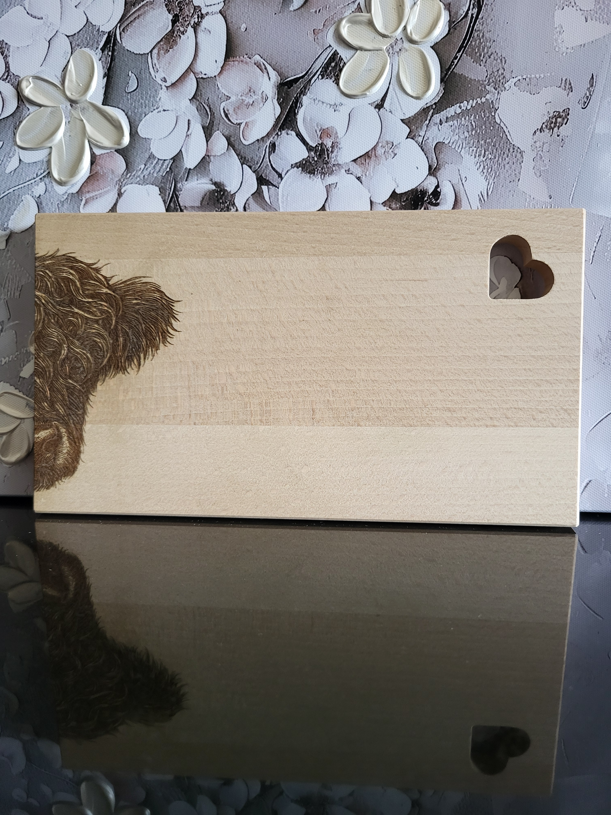 Rechteckiges Schneidebrett aus hellem Buchenholz mit dekorativer Gravur. Auf der linken Seite ist der Kopf eines zotteligen Hochlandrinds detailreich eingraviert, wobei das dichte Fell besonders lebendig wirkt. Rechts oben befindet sich eine ausgestanzte Herzform als praktisches Griffloch, das dem Brett zusätzlich einen charmanten Akzent verleiht. Das massive Holzbrett kombiniert rustikale Natürlichkeit mit liebevollen Details und eignet sich ideal zum Servieren, Schneiden oder als originelles Geschenk.