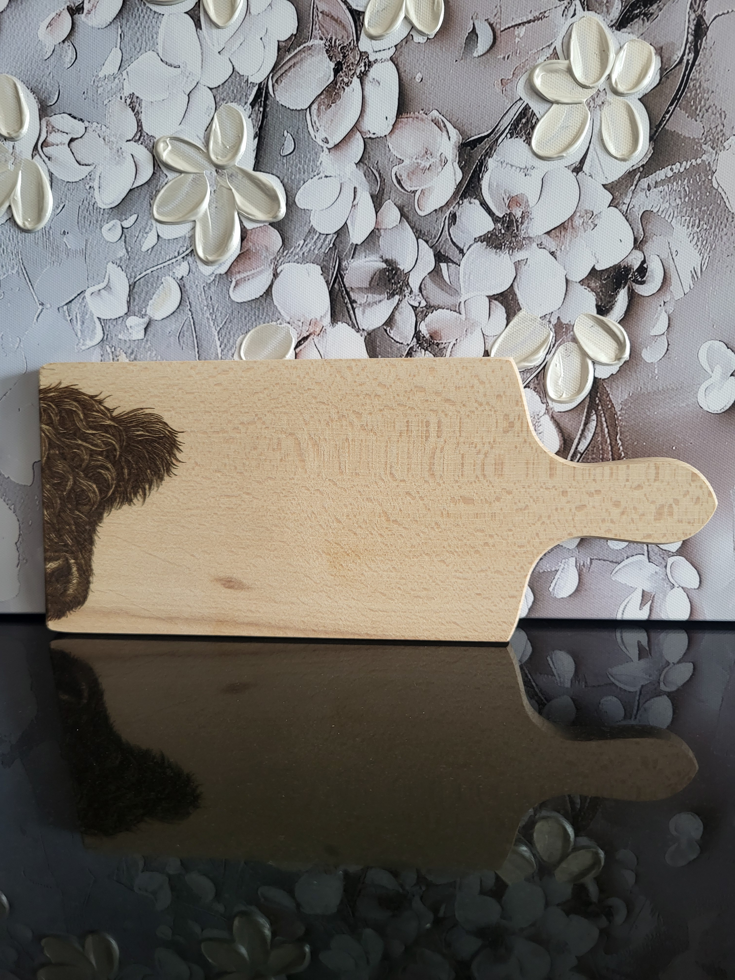 Rechteckiges Schneidebrett aus hellem Buchenholz mit Griff, schlicht und naturbelassen verarbeitet. Auf der rechten unteren Ecke ist detailreich der Kopf eines Hochlandrinds eingraviert, dessen zotteliges Fell besonders plastisch wirkt. Am Griffende befinden sich vier kleine quadratische Einkerbungen als dezentes Design-Element. Das massive Holzbrett vereint Funktionalität mit einem rustikalen, alpinen Charakter und eignet sich ideal zum Schneiden, Servieren oder als Geschenk mit besonderer Gravur.