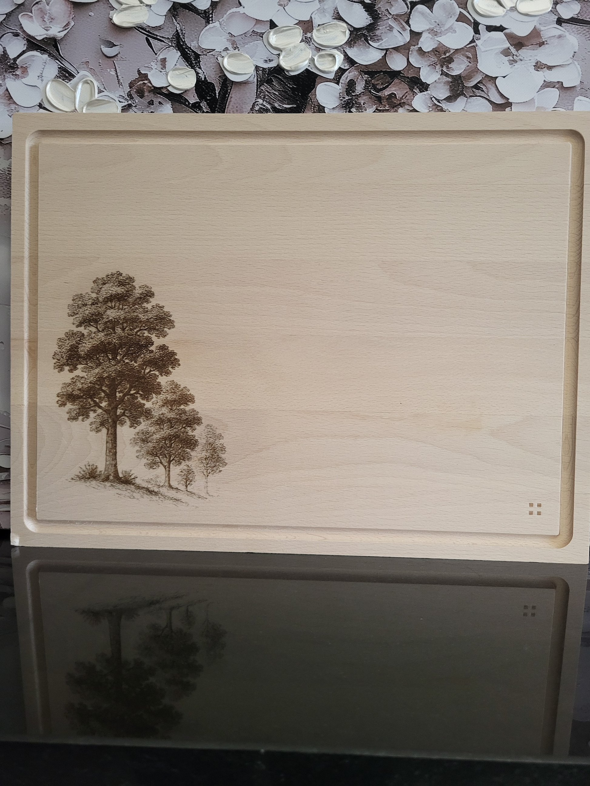 Rechteckiges Schneidebrett aus hellem Buchenholz mit umlaufender Saftrille, schlicht und hochwertig verarbeitet. In der linken unteren Ecke ist eine filigrane Gravur von mehreren Bäumen zu sehen, die detailreich in verschiedenen Größen dargestellt sind und dem Brett eine natürliche, alpine Note verleihen. In der rechten unteren Ecke befindet sich ein dezentes Markenzeichen aus vier kleinen quadratischen Punkten. Das Brett kombiniert Funktionalität mit rustikaler Eleganz und eignet sich sowohl zum Schneiden 
