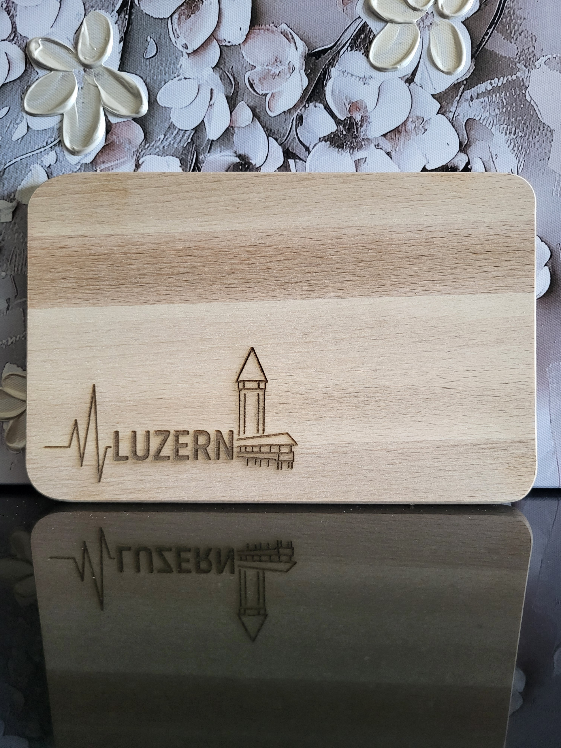 Rechteckiges Schneidebrett aus hellem Buchenholz mit abgerundeten Ecken und schlichter Oberfläche. In der unteren linken Ecke ist eine Gravur mit einem stilisierten Herzschlag-Symbol, dem Schriftzug „LUZERN“ und dem bekannten Wasserturm mit Kapellbrücke zu sehen – ein Wahrzeichen der Stadt Luzern. Das minimalistische Design verbindet traditionelles Schweizer Handwerk mit regionaler Identität und macht das Brett zu einem dekorativen wie funktionalen Küchenaccessoire.