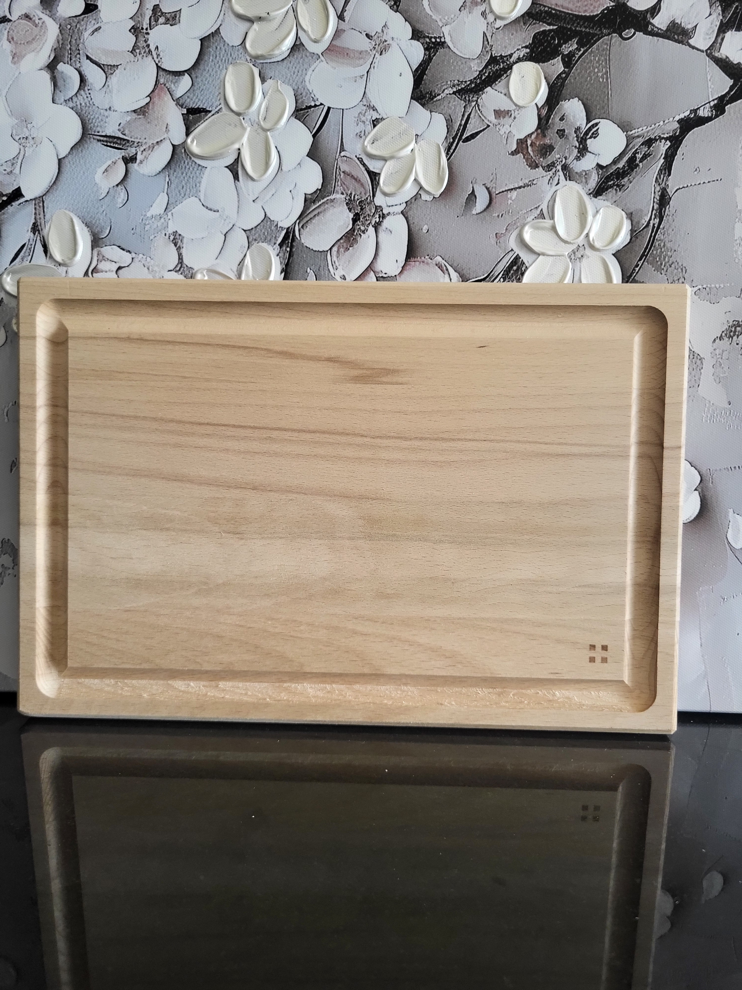 Rechteckiges Schneidebrett aus hellem Holz mit klarer, feiner Maserung. Das Brett besitzt eine umlaufende Saftrille, die Flüssigkeiten auffängt, und überzeugt durch seine schlichte, funktionale Gestaltung. In der unteren rechten Ecke befindet sich ein kleines quadratisches Symbol als feines Detail. Das minimalistische Design macht das Brett vielseitig einsetzbar – ideal zum Schneiden, Servieren oder als stilvolle Basis für Käse- und Wurstplatten.