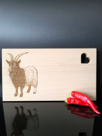 Rechteckiges Schneidebrett aus hellem Buchenholz mit feiner Maserung. Auf der linken Seite ist eine detailreiche Gravur einer Ziege mit langen, geschwungenen Hörnern und zotteligem Fell dargestellt. Auf der rechten oberen Seite befindet sich eine herzförmige Aussparung, die dem Brett einen charmanten Akzent verleiht. Das Brett kombiniert natürliche Schlichtheit mit kunstvoller Gravur – ideal als Küchenaccessoire, Servierbrett oder Geschenk.