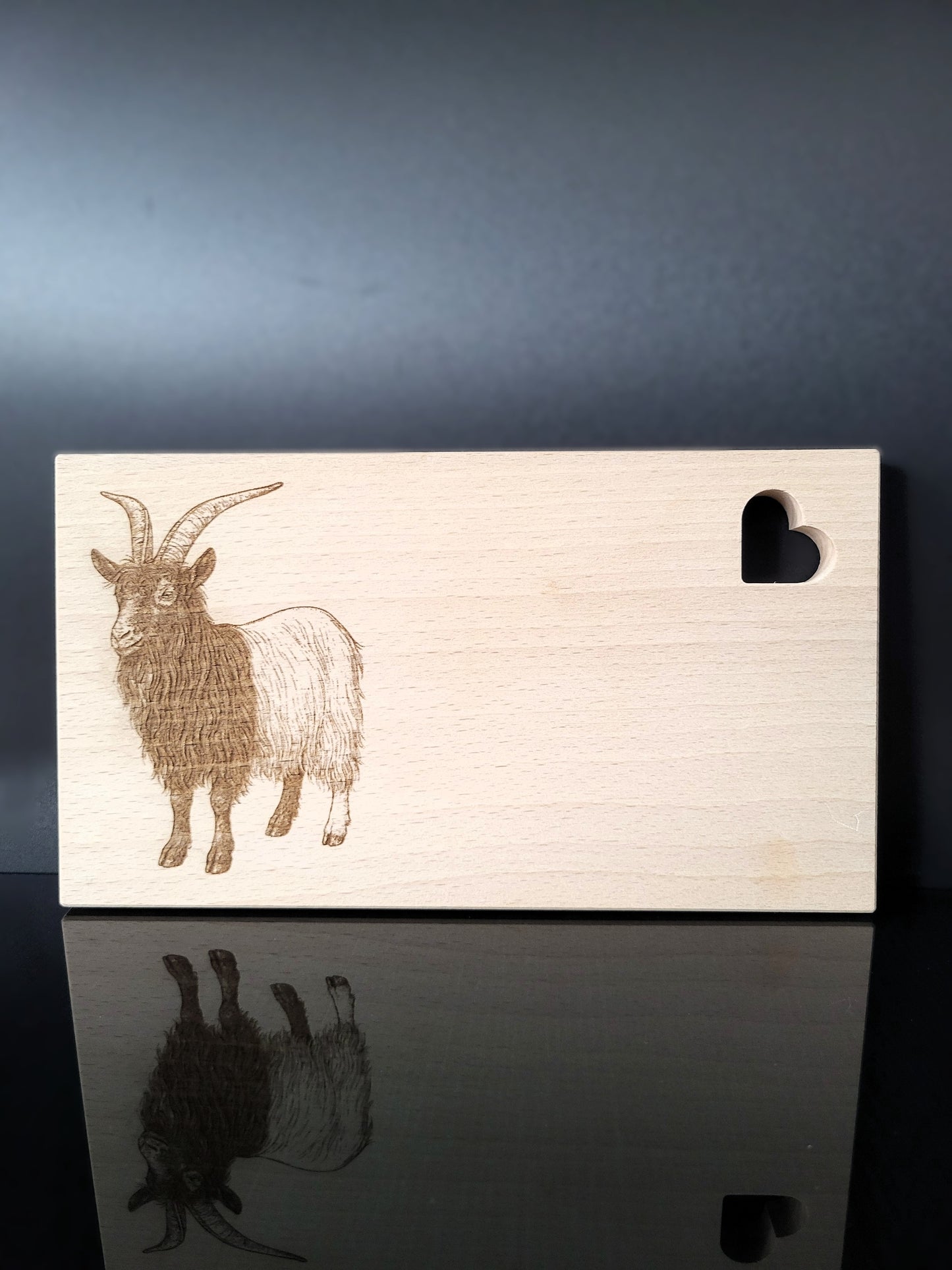 Rechteckiges Schneidebrett aus hellem Buchenholz mit feiner Maserung. Auf der linken Seite ist eine detailreiche Gravur einer Ziege mit langen, geschwungenen Hörnern und zotteligem Fell dargestellt. Auf der rechten oberen Seite befindet sich eine herzförmige Aussparung, die dem Brett einen charmanten Akzent verleiht. Das Brett kombiniert natürliche Schlichtheit mit kunstvoller Gravur – ideal als Küchenaccessoire, Servierbrett oder Geschenk.