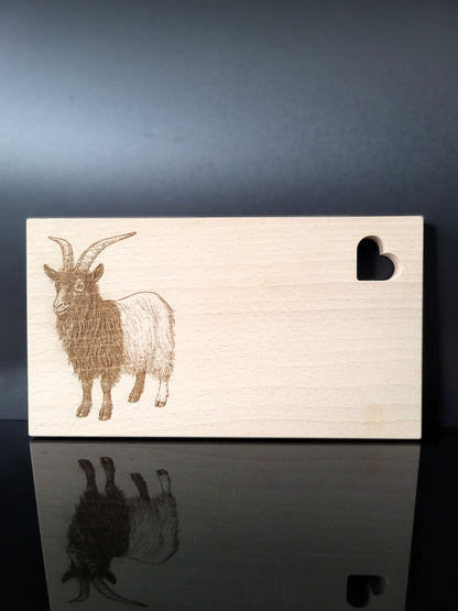Rechteckiges Schneidebrett aus hellem Buchenholz mit feiner Maserung. Auf der linken Seite ist eine detailreiche Gravur einer Ziege mit langen, geschwungenen Hörnern und zotteligem Fell dargestellt. Auf der rechten oberen Seite befindet sich eine herzförmige Aussparung, die dem Brett einen charmanten Akzent verleiht. Das Brett kombiniert natürliche Schlichtheit mit kunstvoller Gravur – ideal als Küchenaccessoire, Servierbrett oder Geschenk.