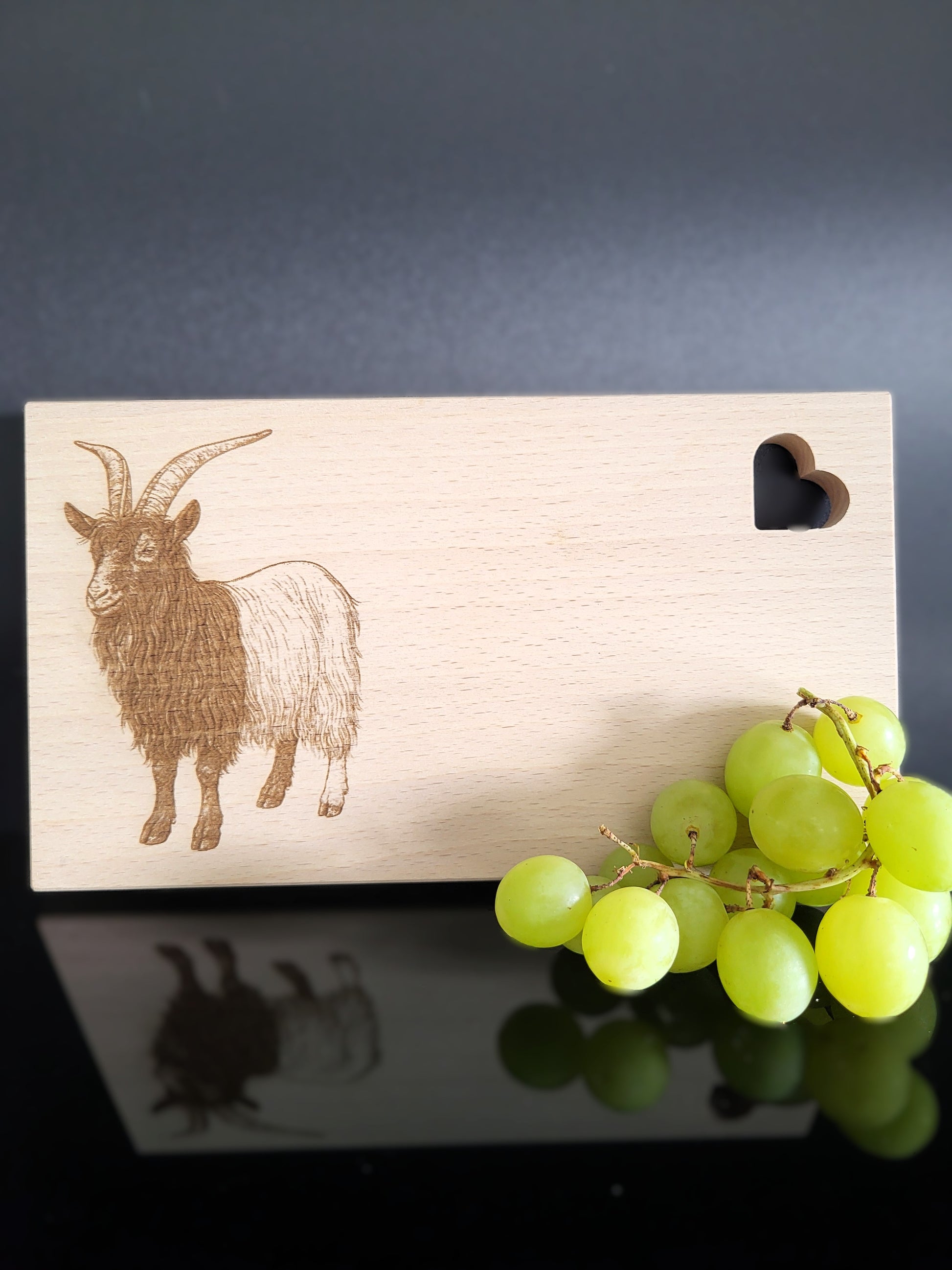 Rechteckiges Schneidebrett aus hellem Buchenholz mit feiner Maserung. Auf der linken Seite ist eine detailreiche Gravur einer Ziege mit langen, geschwungenen Hörnern und zotteligem Fell dargestellt. Auf der rechten oberen Seite befindet sich eine herzförmige Aussparung, die dem Brett einen charmanten Akzent verleiht. Das Brett kombiniert natürliche Schlichtheit mit kunstvoller Gravur – ideal als Küchenaccessoire, Servierbrett oder Geschenk.