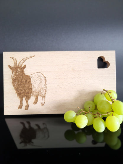 Rechteckiges Schneidebrett aus hellem Buchenholz mit feiner Maserung. Auf der linken Seite ist eine detailreiche Gravur einer Ziege mit langen, geschwungenen Hörnern und zotteligem Fell dargestellt. Auf der rechten oberen Seite befindet sich eine herzförmige Aussparung, die dem Brett einen charmanten Akzent verleiht. Das Brett kombiniert natürliche Schlichtheit mit kunstvoller Gravur – ideal als Küchenaccessoire, Servierbrett oder Geschenk.