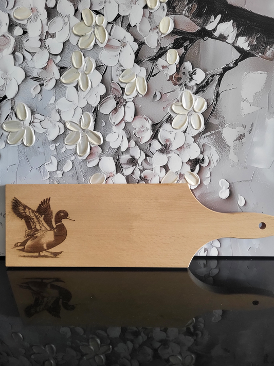 Rechteckiges Schneidebrett aus hellem Buchenholz mit Griff und Loch zum Aufhängen. Auf der linken Seite ist eine detaillierte Gravur einer Ente zu sehen, die mit ausgebreiteten Flügeln im Begriff ist, zu landen oder loszufliegen. Die feine Gravur hebt die Struktur der Federn und den lebendigen Ausdruck hervor. Das Brett kombiniert praktische Funktionalität mit dekorativer Tiergravur und eignet sich sowohl als Küchenutensil als auch als originelles Geschenk.