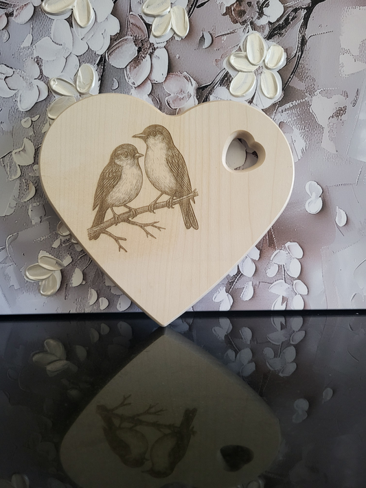 Herzförmiges Schneidebrett aus hellem Holz mit feiner Gravur von zwei Vögeln, die nebeneinander auf einem Ast sitzen. Die Vögel sind detailreich gearbeitet und vermitteln ein romantisches, naturverbundenes Motiv. Oben rechts befindet sich ein ausgestanztes Herzloch, das als Griff oder Aufhängung dient. Das Schneidebrett vereint liebevolles Design mit praktischer Funktion und eignet sich sowohl als Küchenaccessoire als auch als dekoratives Geschenk.