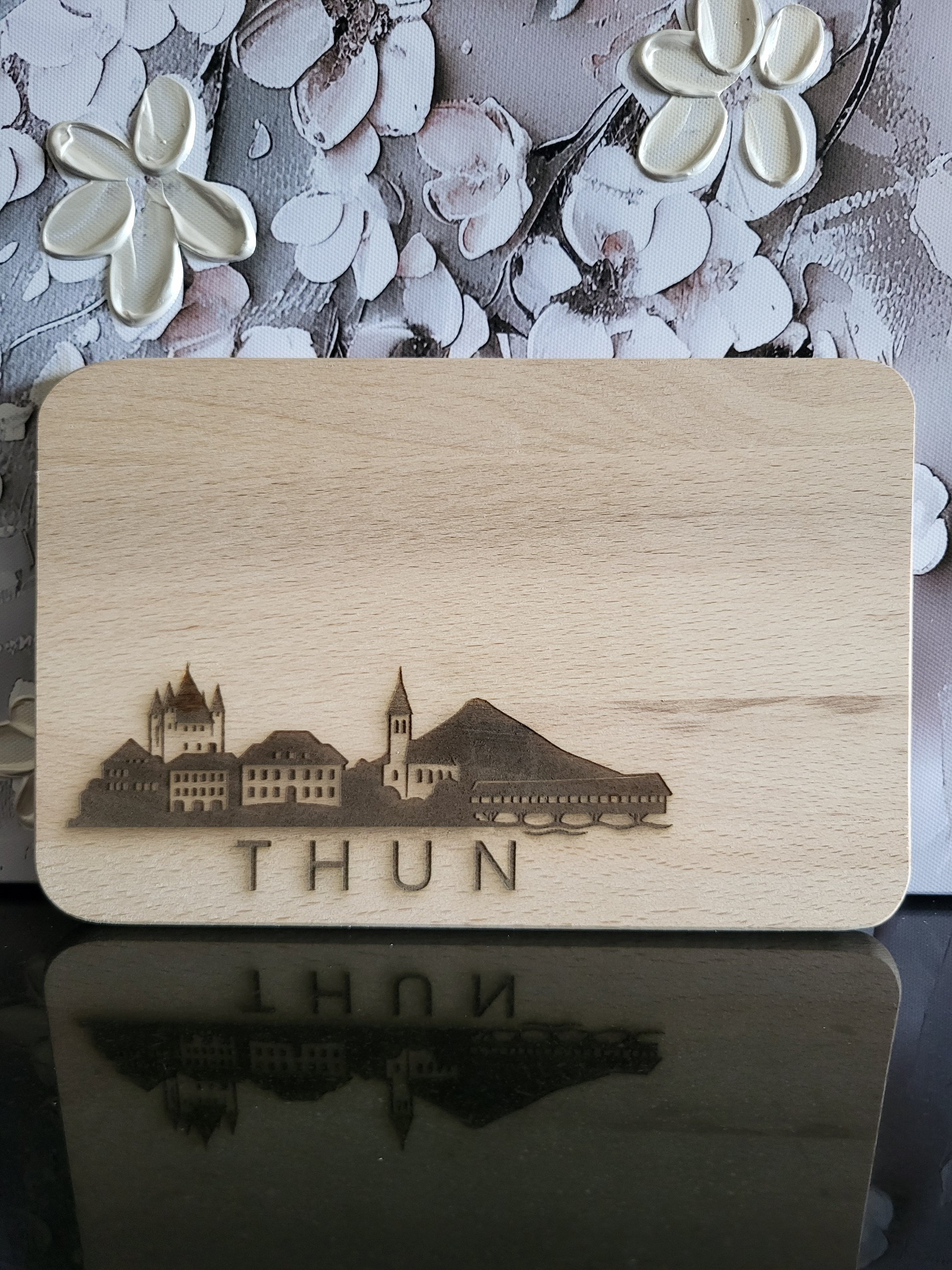 Rechteckiges Schneidebrett aus hellem Buchenholz mit abgerundeten Ecken. Auf der Vorderseite ist eine feine Gravur der Skyline von Thun zu sehen: Links das markante Schloss Thun mit seinen Türmen, daneben historische Gebäude und eine Kirche mit spitzem Turm. Rechts schließt sich ein stilisierter Berg sowie die überdachte Holzbrücke über die Aare an. Unterhalb der Gravur steht der Schriftzug „THUN“ in Großbuchstaben. Das Brett verbindet praktisches Küchenutensil mit einem dekorativen Motiv, das die Stadt Thu