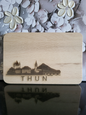 Rechteckiges Schneidebrett aus hellem Buchenholz mit abgerundeten Ecken. Auf der Vorderseite ist eine feine Gravur der Skyline von Thun zu sehen: Links das markante Schloss Thun mit seinen Türmen, daneben historische Gebäude und eine Kirche mit spitzem Turm. Rechts schließt sich ein stilisierter Berg sowie die überdachte Holzbrücke über die Aare an. Unterhalb der Gravur steht der Schriftzug „THUN“ in Großbuchstaben. Das Brett verbindet praktisches Küchenutensil mit einem dekorativen Motiv, das die Stadt Thu
