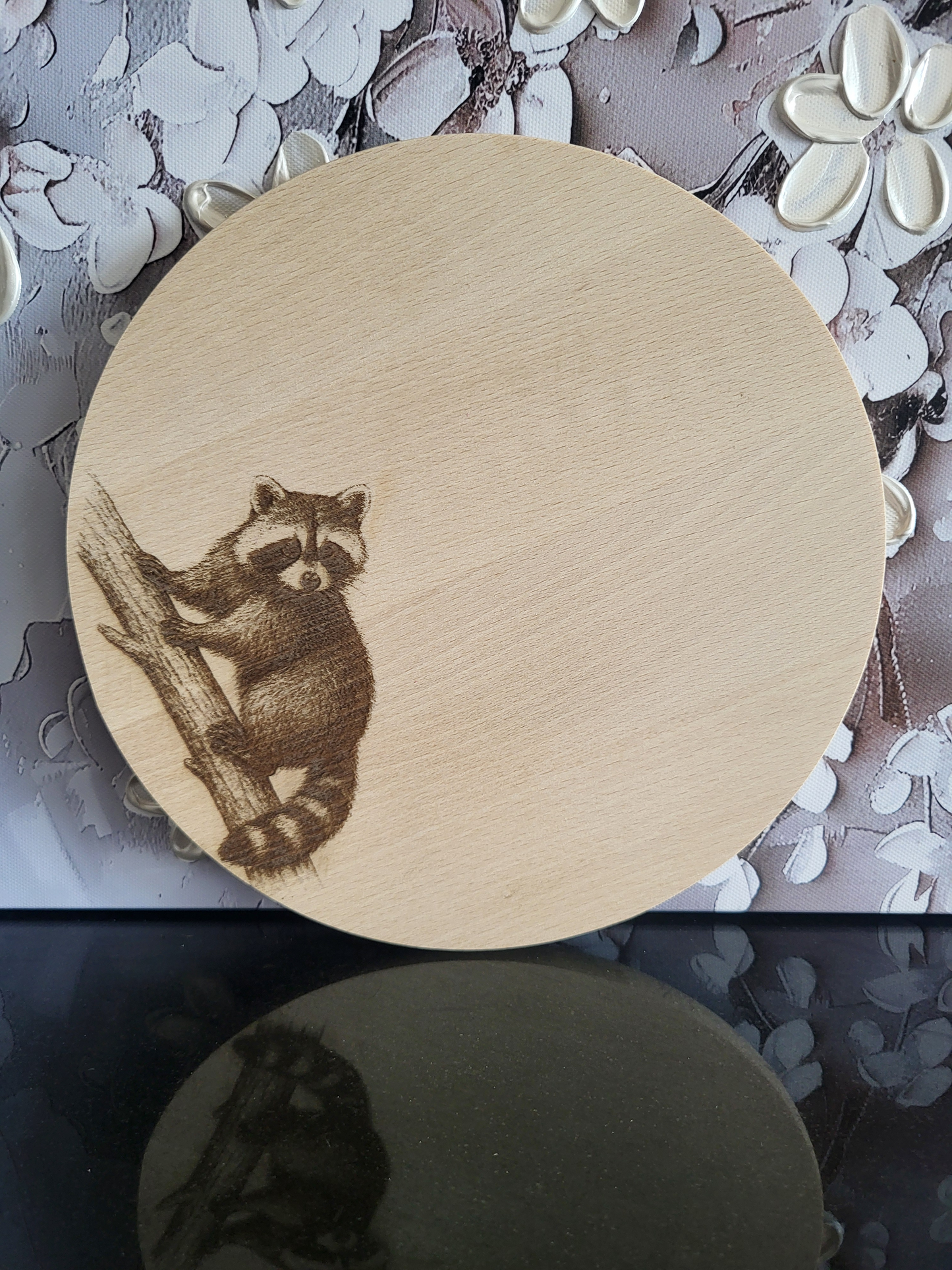 Rundes Schneidebrett aus hellem Holz mit einer feinen Gravur eines Waschbären. Der Waschbär sitzt auf einem Baumstamm und blickt aufmerksam nach vorne. Die Gravur ist detailreich gearbeitet und hebt sich kontrastreich vom Holz ab. Das Brett verbindet Naturmotiv mit praktischer Funktion und eignet sich ideal als Küchenaccessoire oder Geschenk mit besonderem Charme.