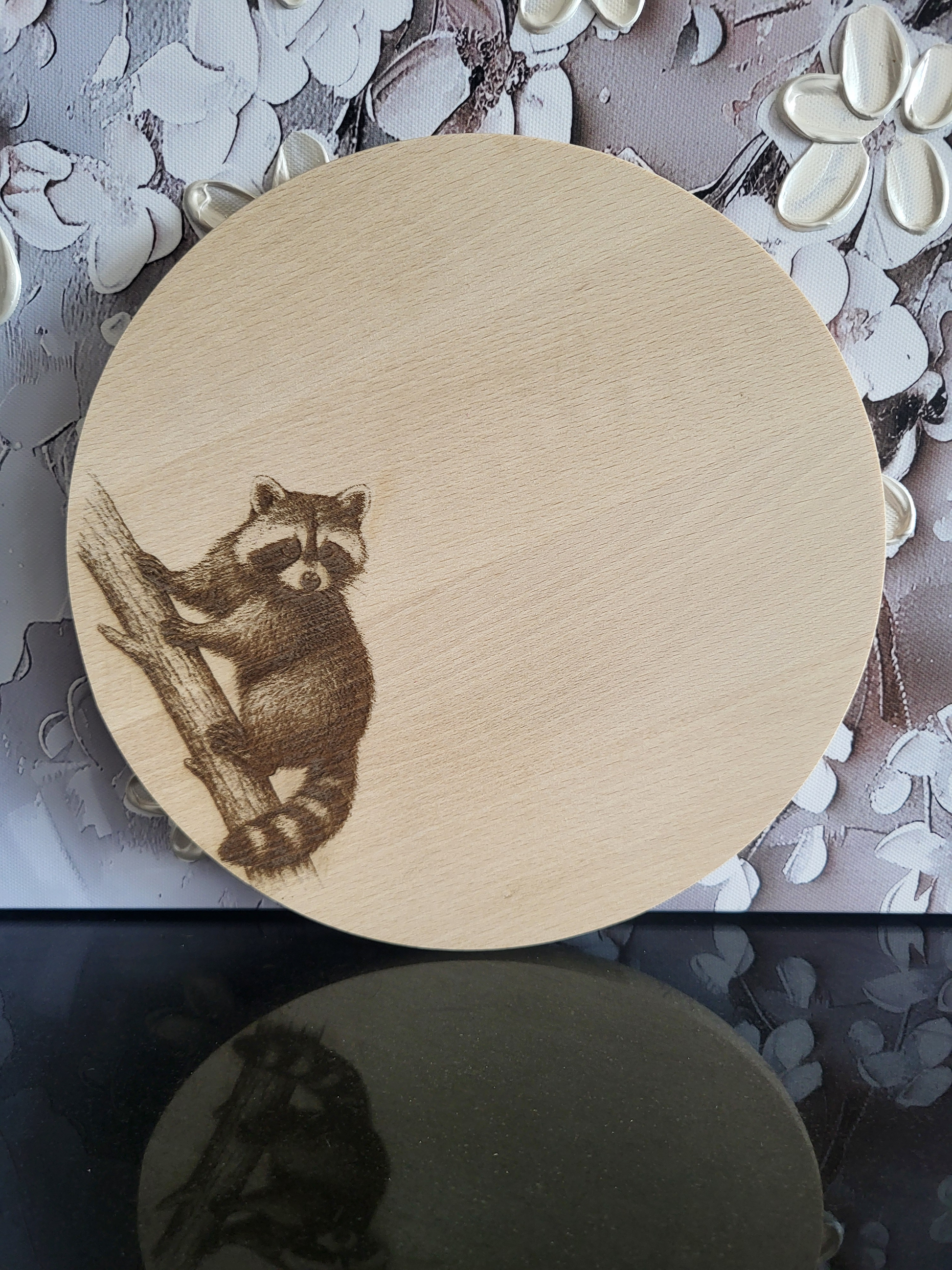 Rundes Schneidebrett aus hellem Holz mit einer feinen Gravur eines Waschbären. Der Waschbär sitzt auf einem Baumstamm und blickt aufmerksam nach vorne. Die Gravur ist detailreich gearbeitet und hebt sich kontrastreich vom Holz ab. Das Brett verbindet Naturmotiv mit praktischer Funktion und eignet sich ideal als Küchenaccessoire oder Geschenk mit besonderem Charme.