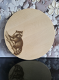 Rundes Schneidebrett aus hellem Holz mit einer feinen Gravur eines Waschbären. Der Waschbär sitzt auf einem Baumstamm und blickt aufmerksam nach vorne. Die Gravur ist detailreich gearbeitet und hebt sich kontrastreich vom Holz ab. Das Brett verbindet Naturmotiv mit praktischer Funktion und eignet sich ideal als Küchenaccessoire oder Geschenk mit besonderem Charme.