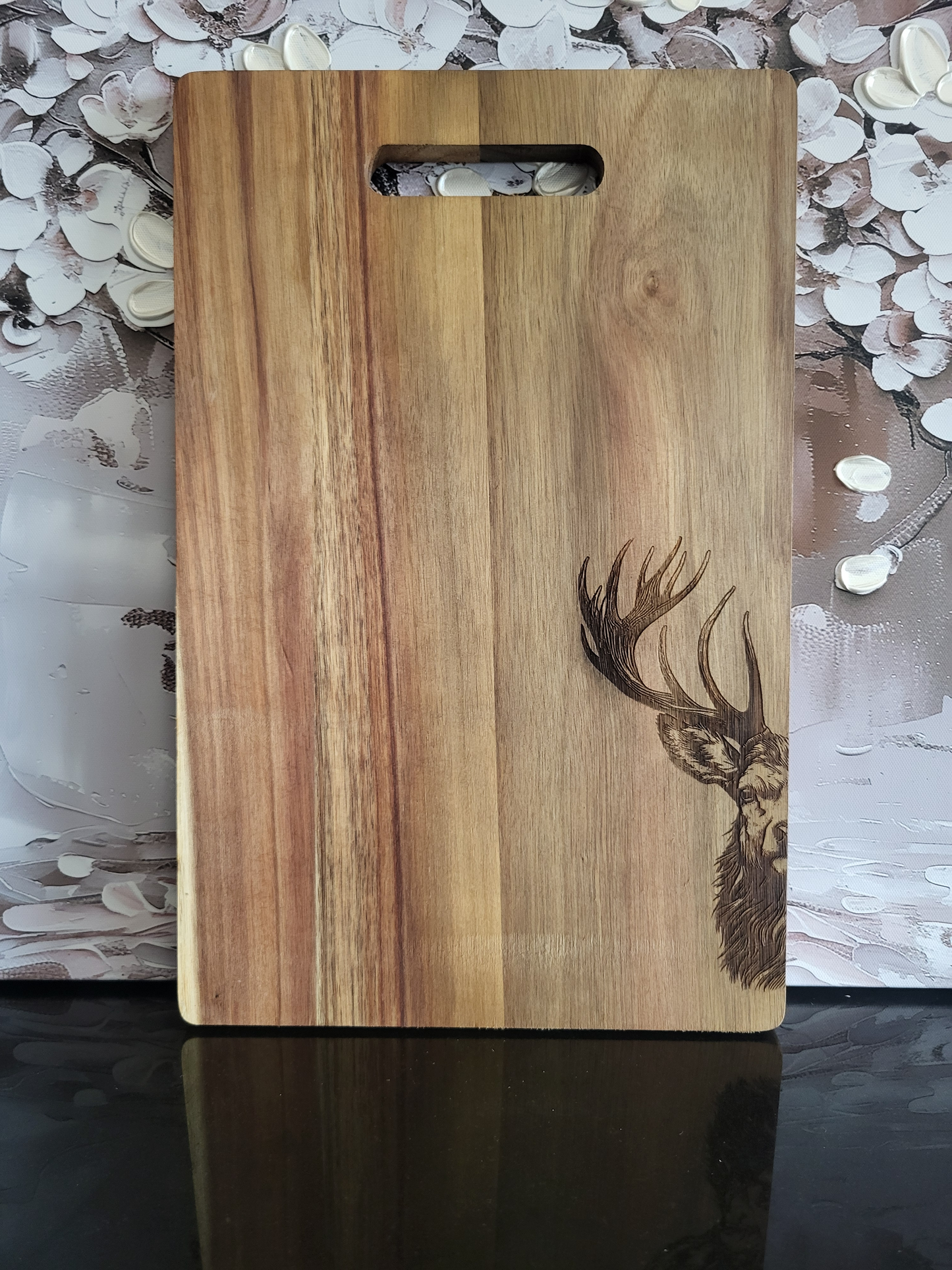 Rechteckiges Schneidebrett aus Akazienholz mit natürlicher Maserung und Grifföffnung im oberen Bereich. In der unteren rechten Ecke befindet sich eine detailreiche Gravur eines Hirschkopfes mit prächtigem Geweih. Das edle Design kombiniert rustikale Holzoptik mit feiner Gravurkunst und macht das Brett sowohl funktional als auch dekorativ – ideal als Küchenutensil oder Geschenk mit Alpencharme.