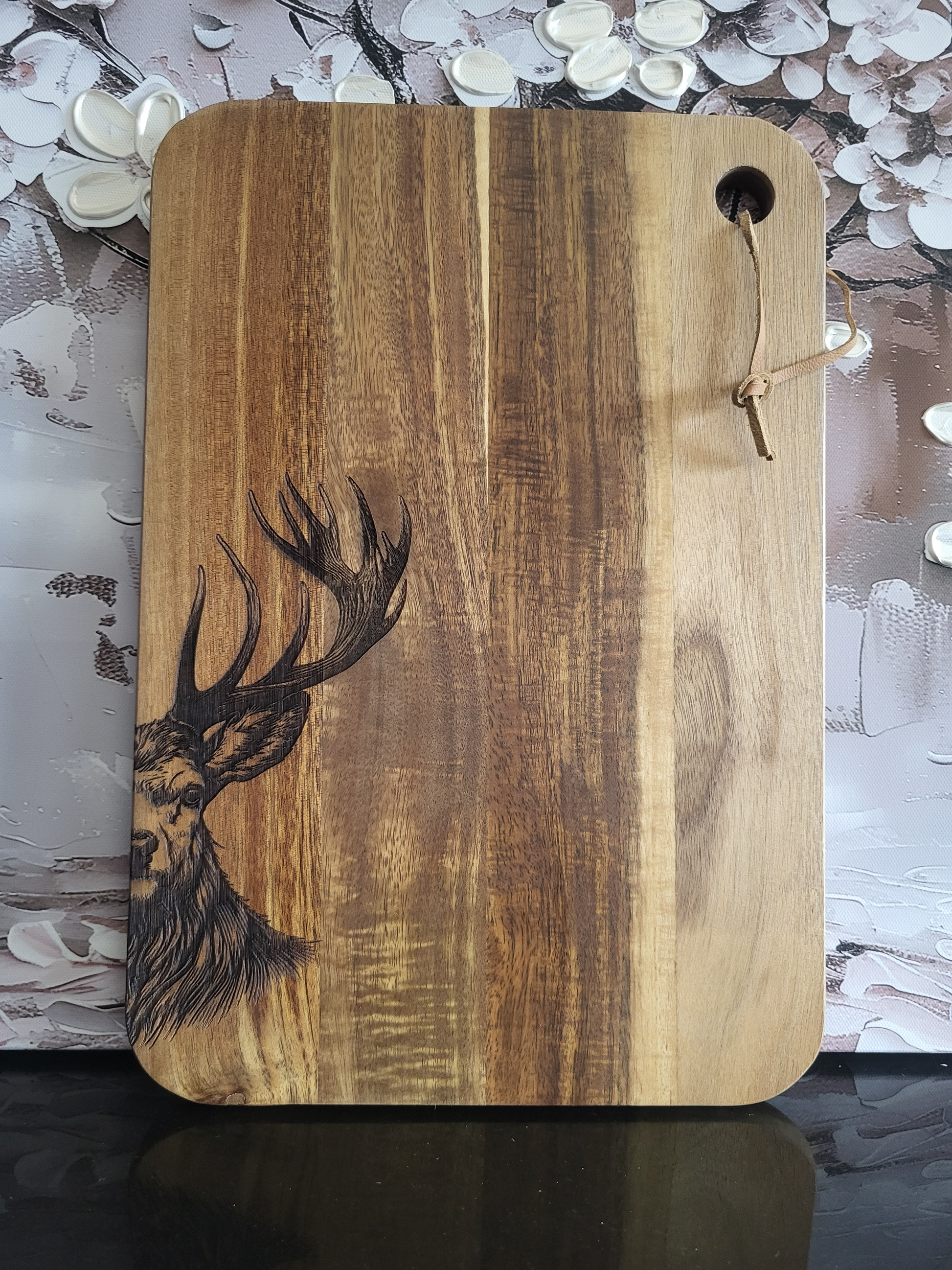 Rechteckiges Schneidebrett aus massivem Akazienholz mit markanter Maserung. In der unteren linken Ecke ist ein detailreich gravierter Hirschkopf mit großem Geweih dargestellt. Oben rechts befindet sich eine runde Aufhängeöffnung mit einer Lederschnur zum praktischen Verstauen. Das Brett vereint rustikales Naturdesign mit kunstvoller Gravur und eignet sich ideal zum Schneiden, Servieren oder als dekoratives Geschenkstück.