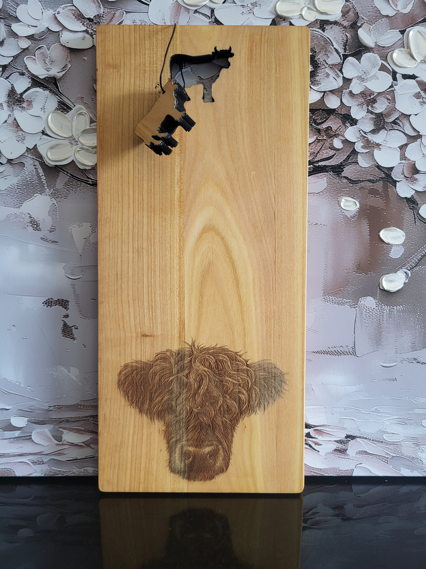 Rechteckiges Schneidebrett aus hellem Kirschholz mit natürlicher Maserung. Unten ist eine detailreiche Gravur des Kopfes einer Kuh mit lockigem Fell dargestellt, die das Brett schmückt. Oben befindet sich ein ausgestanztes Kuh-Motiv, aus dem ein kleiner Anhänger in Kuhform hängt. Das Brett vereint rustikales Naturdesign mit liebevoller Gravur und traditionellem Charme und eignet sich ideal zum Schneiden, Servieren oder als dekoratives Geschenkstück.