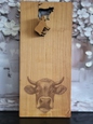 Längliches Schneidebrett aus hellem Holz mit natürlicher Maserung. Am unteren Rand ist detailreich der Kopf einer Kuh mit Hörnern und feiner Fellstruktur eingraviert. Im oberen Bereich befindet sich ein ausgestanztes Kuh-Motiv, aus dem ein kleiner Holzanhänger in Kuhform herabhängt. Das Brett verbindet rustikale Handwerkskunst mit praktischer Funktion und dekorativem Charme.