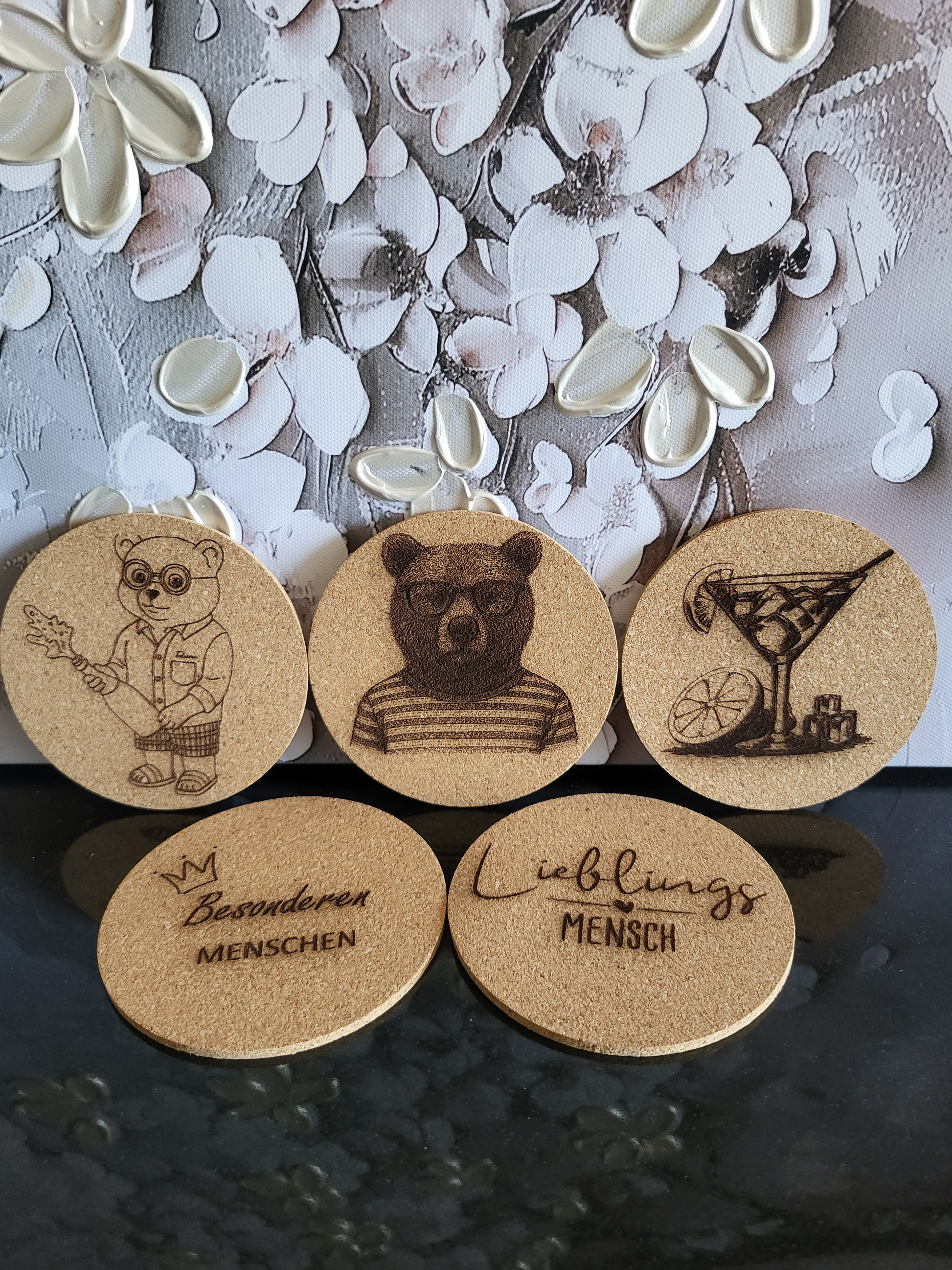 Set aus fünf Korkuntersetzern für Apéro oder Cocktails. Die gravierten Untersetzer zeigen verschiedene Motive wie einen Bären mit Brille, einen Bären im Ringelshirt und ein Martini-Glas. Zwei weitere Untersetzer sind mit den Sprüchen "Lieblingsmensch" und "Besonderen MENSCHEN" beschriftet. Perfekt als Geschenkset für Liebhaber von stilvollem Bar-Zubehör oder als originelles Geschenk für Freunde und Familie.