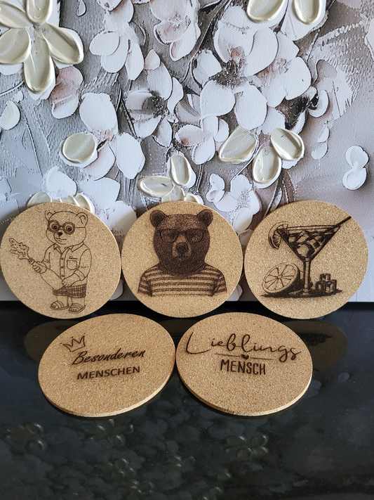 Set aus fünf Korkuntersetzern für Apéro oder Cocktails. Die gravierten Untersetzer zeigen verschiedene Motive wie einen Bären mit Brille, einen Bären im Ringelshirt und ein Martini-Glas. Zwei weitere Untersetzer sind mit den Sprüchen "Lieblingsmensch" und "Besonderen MENSCHEN" beschriftet. Perfekt als Geschenkset für Liebhaber von stilvollem Bar-Zubehör oder als originelles Geschenk für Freunde und Familie.
