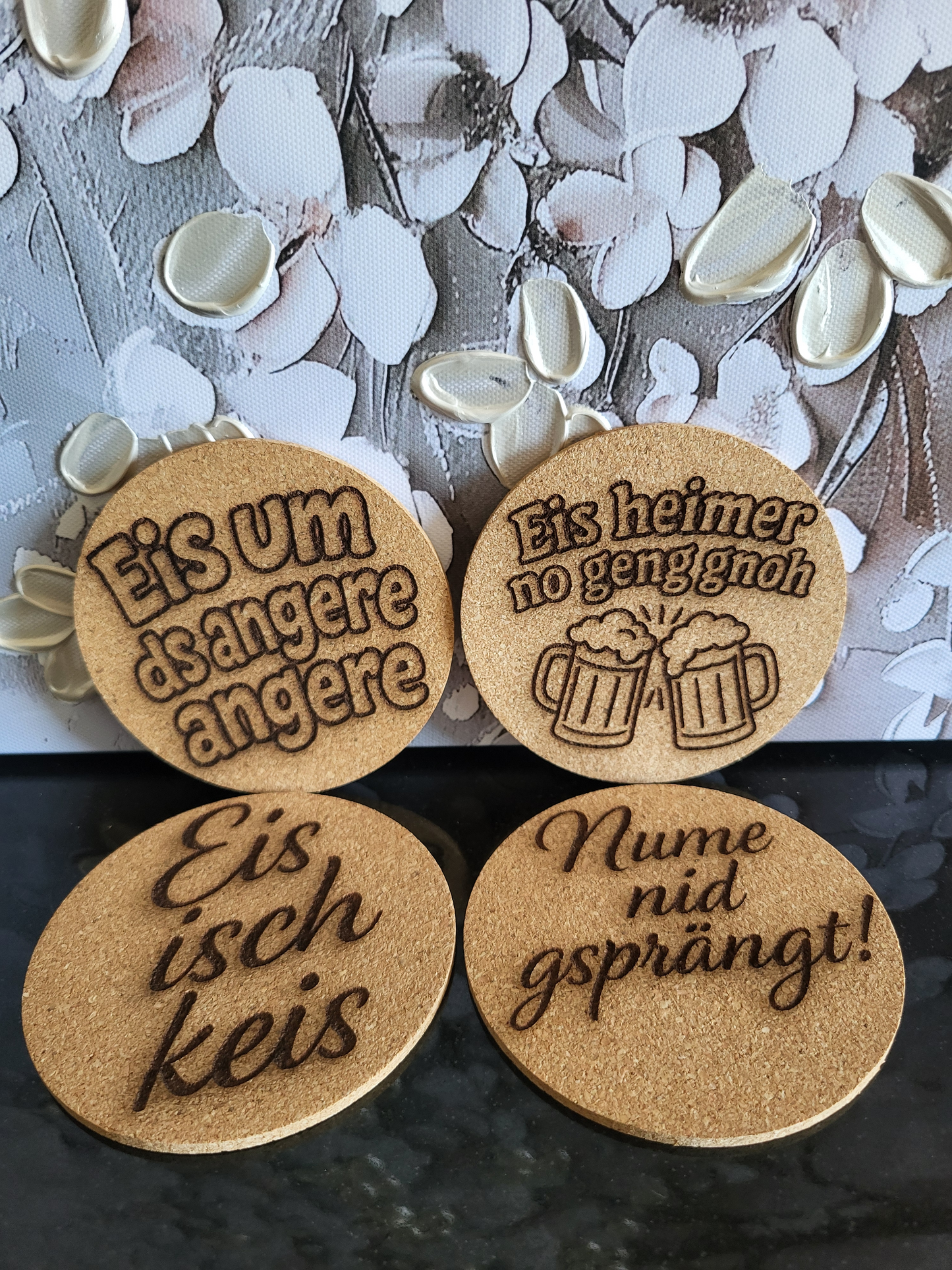 Set aus vier runden Korkuntersetzern mit Berndeutsch-Sprüchen. Die gravierten Sprüche sind: "Eis um ds andere angere", "Eis heimer no geng gnoh" (mit zwei Bierkrügen), "Eis isch keis" und "Nume nid gsprängt!". Perfekt als Geschenkidee für Freunde des Berner Dialekts und als humorvolles Bar-Zubehör.