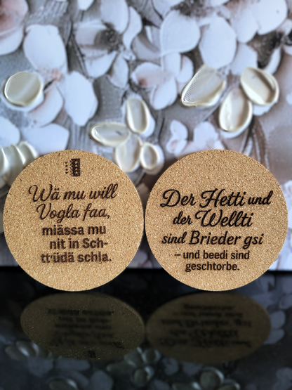 Zwei handgefertigte Kork-Untersetzer von Alpcut mit lasergravierten, humorvollen Sprüchen im Walliser Dialekt, darunter "Wä mu will Vogla faa, miässa mu nit in Schtrüdä schla." und "Der Hetti und der Wellti sind Brieder gsi – und beedi sind gschtorbe.". Perfekte Geschenkidee mit Schweizer Charme.