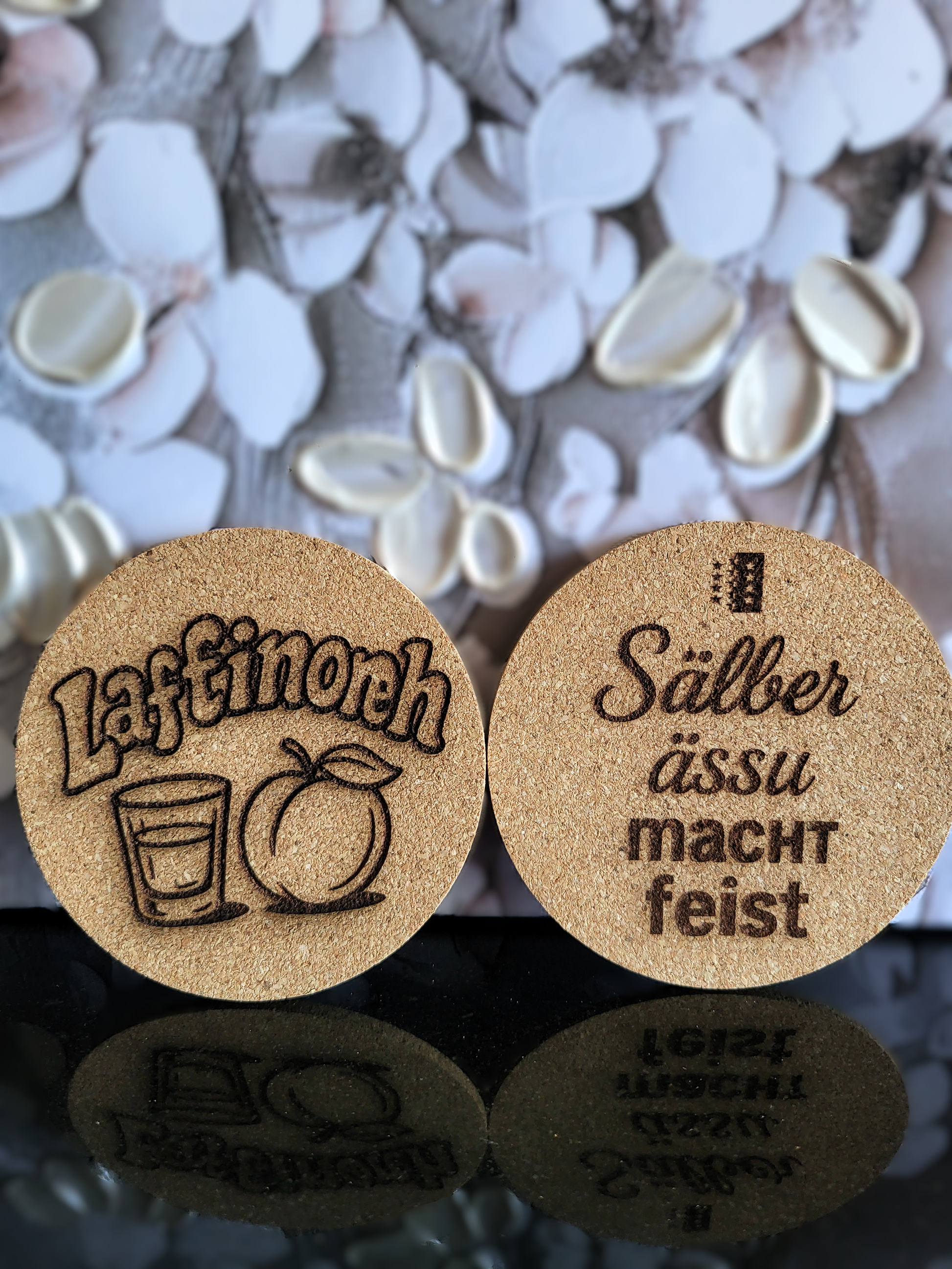 Handgefertigte Kork-Untersetzer von Alpcut, lasergraviert mit Motiven und humorvollen Sprüchen im Walliser Dialekt wie "Läftinörh" mit Aprikose und Glas, sowie "Sälber ässu macht feist".